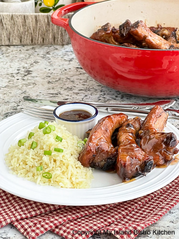 Spareribs in sweet sauce alongside steamed rice on white plate. Red Dutch Oven containing spareribs in background