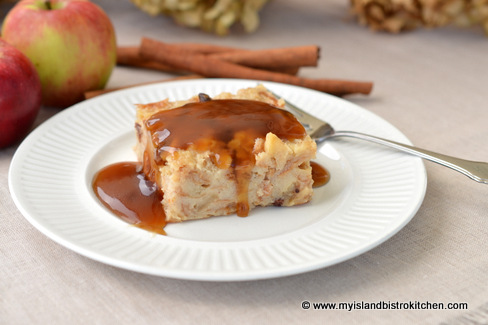 Slice of Apple Bread Pudding on plate. Pudding is drizzled with Maple Sauce. Apples and cinnamon sticks appear in the background.