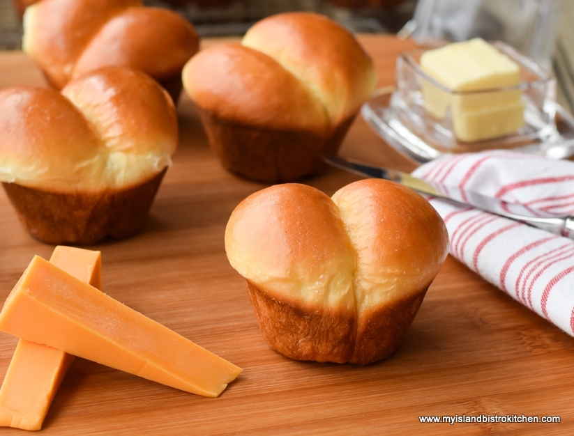 Puffy golden-brown dinner rolls on a bread board alongside cheddar cheese, butter, and a red and white striped tea towel