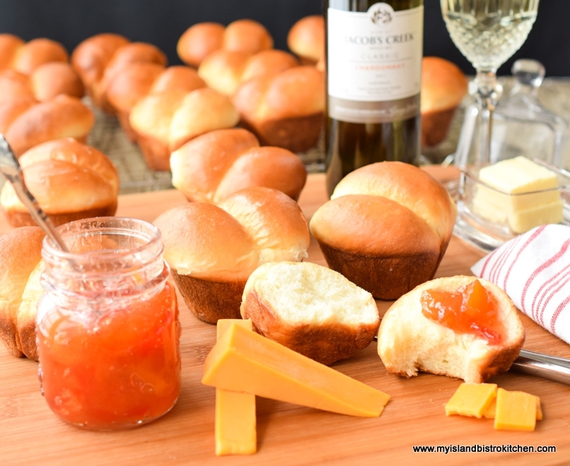 Lots of dinner rolls with butter, cheese, peach jam, and wine on a bread board