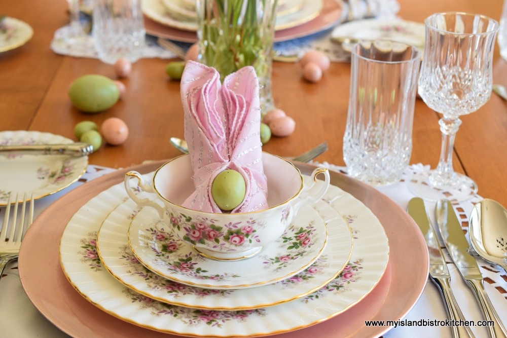 Close-up of placesetting featuring dinner plate, salad plate, and cream soup bowl. Dinnerware is white background with small pink and purple flower border. Pink napkin folded into the shape of bunny ears is placed inside the soup bowl