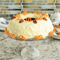 Cream Cheese frosted Carrot Cake sits on pedestal glass cake plate on a counter