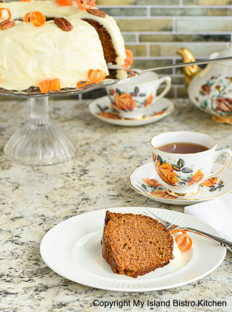 Slice of Carrot Cake on a dessert plate with Carrot Cake on pedestal glass cake plate in the background. Two pretty teacups and a floral teapot are also in the background