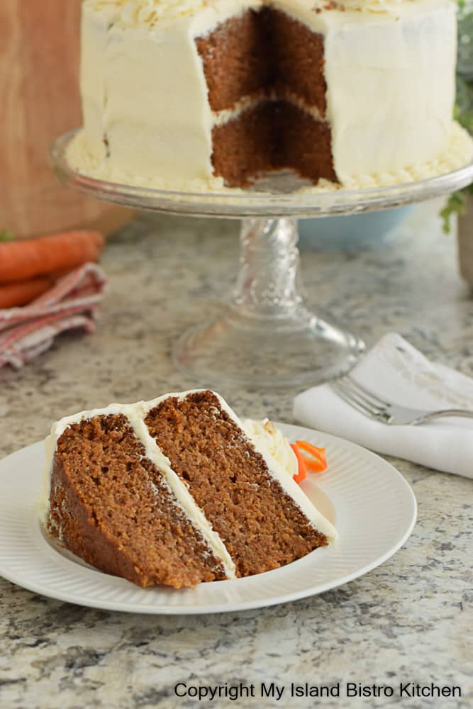 Slice of two-layer Carrot Cake sits on dessert plate with the cake sitting on a pedestal glass cake plate in the background