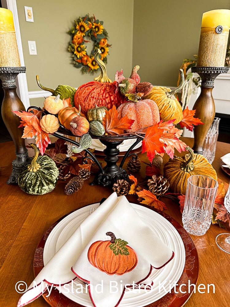 A pretty ivory napkin with an orange pumpkin motif sits atop the dinner plate at each place setting. A colorful table centerpiece of multiple pumpkins in different colors and textures is in the background.