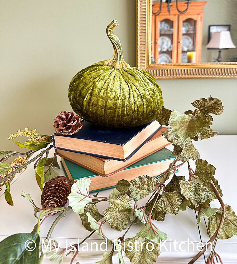 A fireplace vignette comprised of a green velvet pumpkin on a stack of books surrounded by some greenery and pinecones