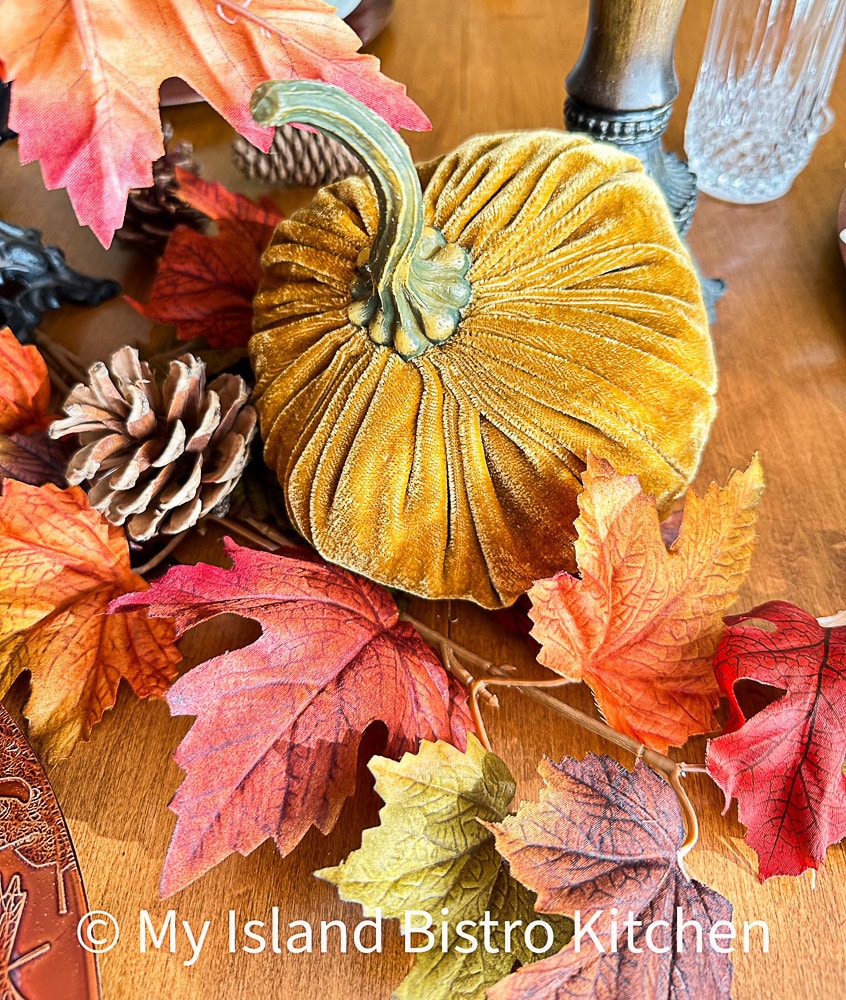 A gold velvet pumpkin sits amidst colored leaves and pinecones on a table