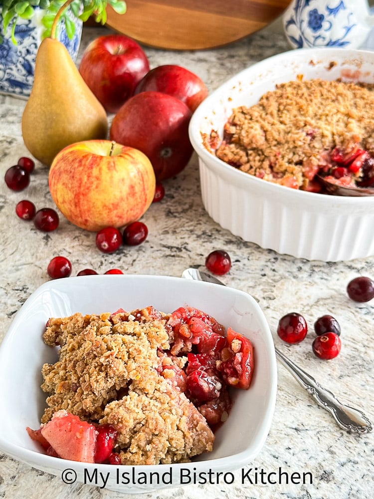 Bowl of Fruit Crisp in foreground; Baking Pan of Crisp surrounded by Apples, Pear, and Cranberries in Background