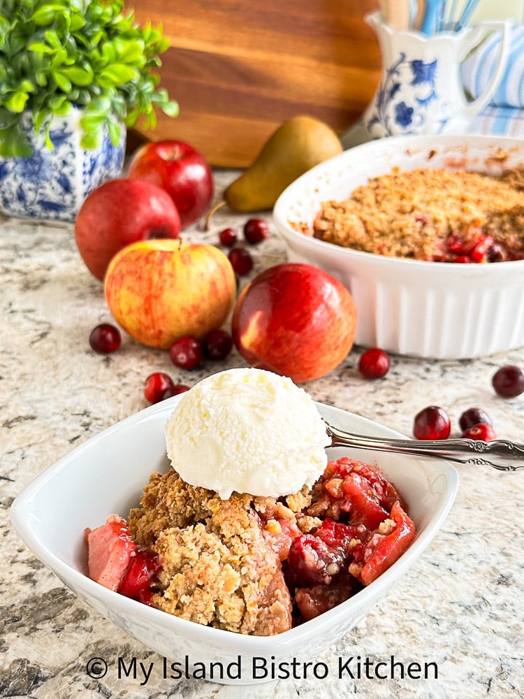 Bowl of fruit crisp topped with vanilla icecream in foreground with apples, pears, and cranberries in background