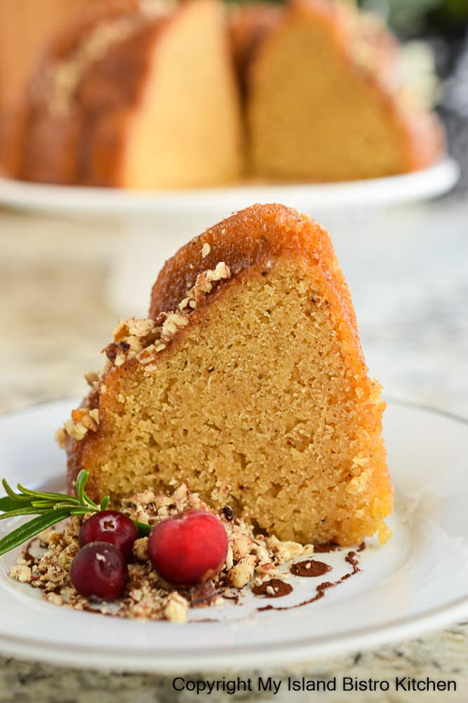 Slice of Rum Cake on plate decorated with chopped nuts, cranberries, and a sprig of Rosemary