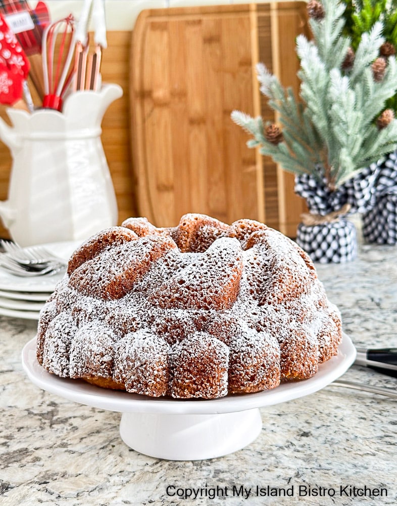 Icing sugar dusted Rum Cake sits on white pedestal cake plate on counter