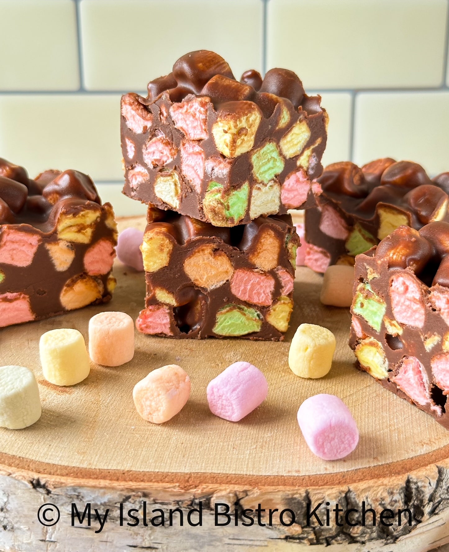 Pieces of Confetti Square sit atop a wooden board on counter with colorful miniature marshmallows surrounding them.
