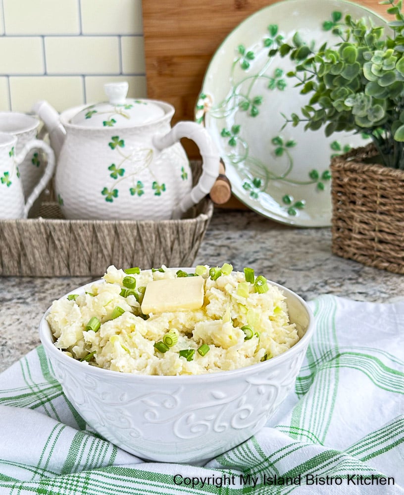 Bowl of Irish Mash sits, surrounded by a green and white tea towel, on counter in front of a Shamrock Teapot and Plate in the background