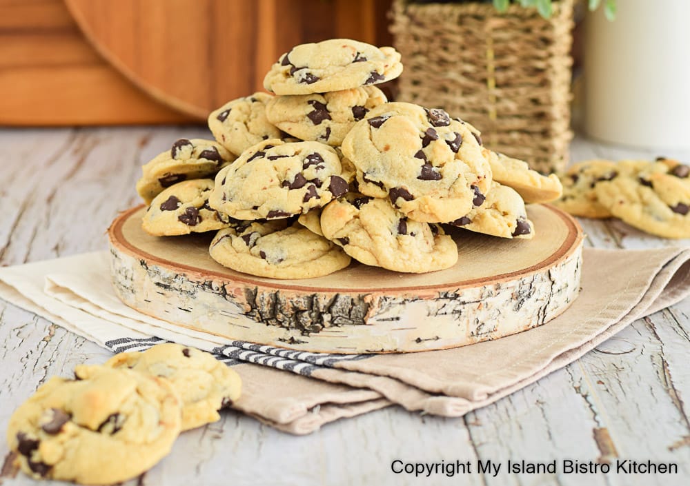 A tower of homemade cookies filled with chocolate chips sits atop a piece of wood on countertop