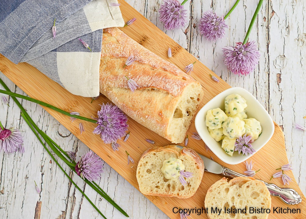 Baguette on breadboard along with a small bowl of compound butter pats and chive blossoms