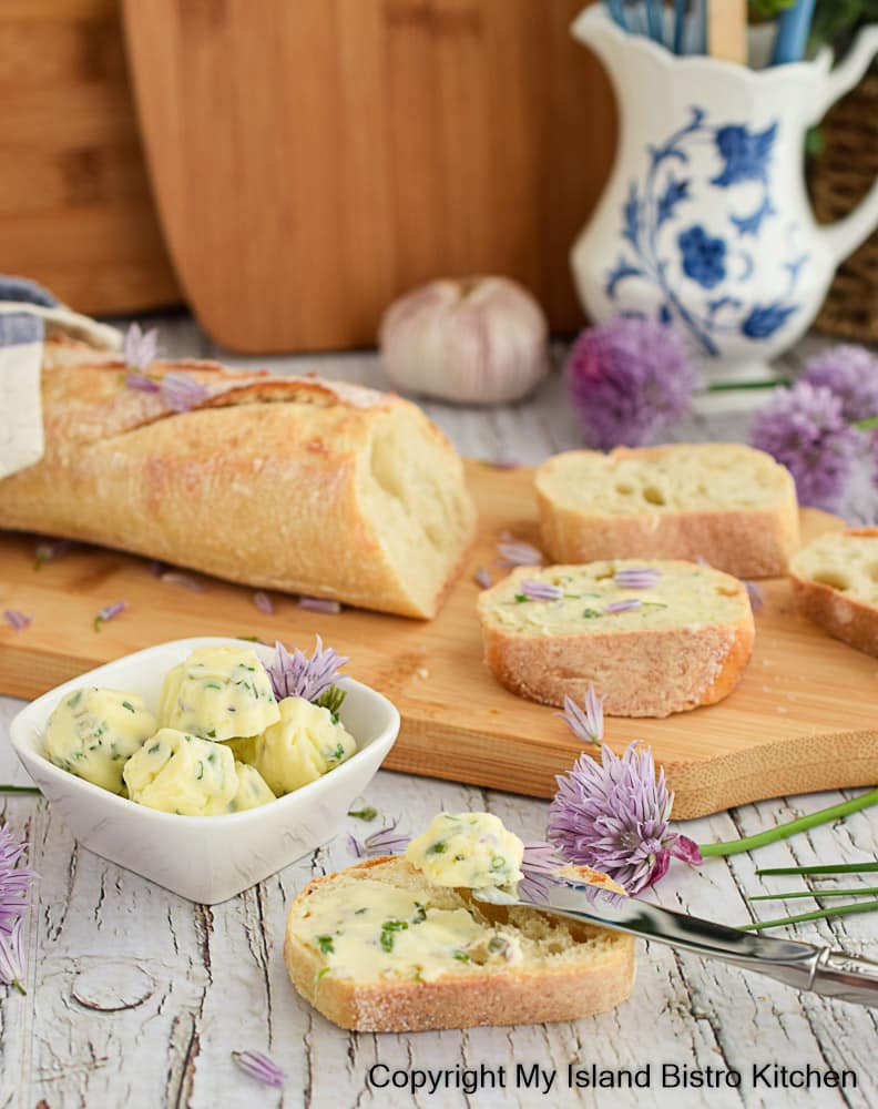 Sliced baguette on wooded bread board on counter surrounded by fresh chive blossoms and a small bowl of compound butter pats