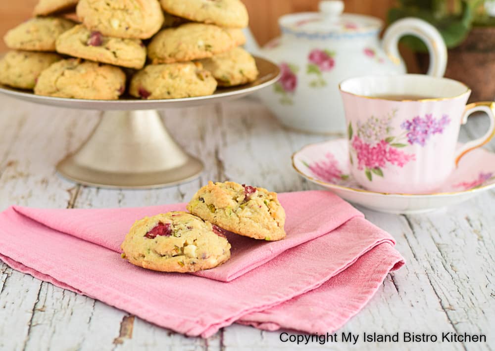 Two Rhubarb Cookies on a pink napkin with a pedestal plate of cookies in the background alongside a pretty pink teacup and white floral teapot