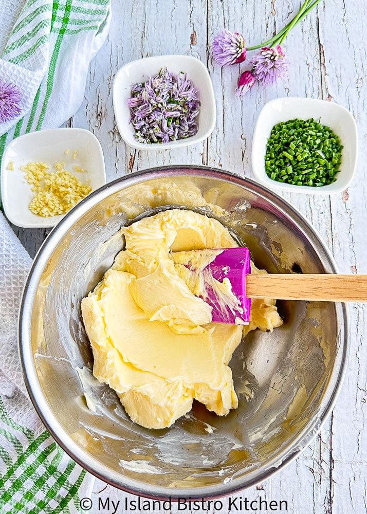 Mixing compound butter in bowl with small bowls of chopped garlic, onion chive blossoms, and chopped chives in background