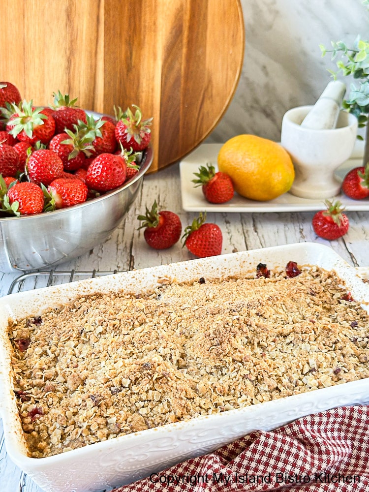 Baked fruit crisp in white casserole sits on counter with bowl of strawberries in background