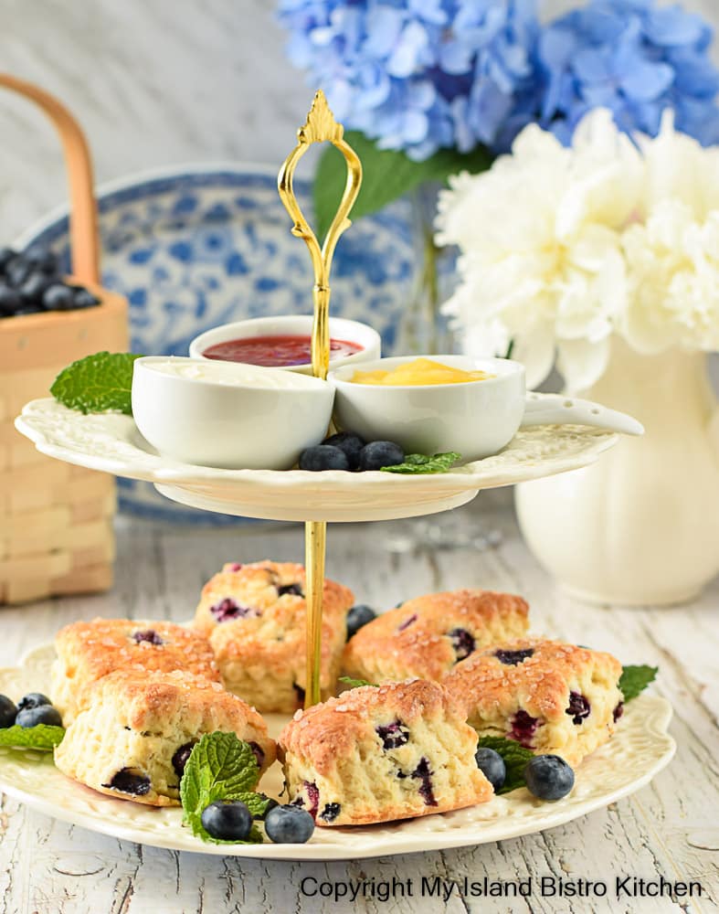 Two-tiered server containing scones and spreads (lemon curd, jam, and English double cream). A basket of blueberries, a blue plate, and bouquets of blue hydrangea and white peonies are in the background.