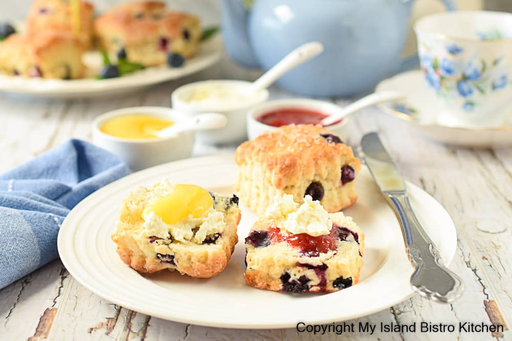 Scones topped with lemon curd, jam, and English double cream sit on white plate surrounded by small dishes of the scone spreads