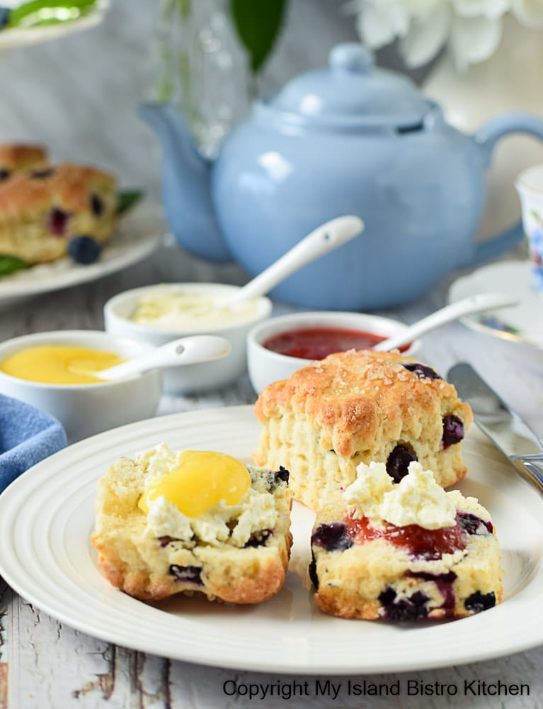 Scones on a white plate with jam, lemon curd, and English double cream with a blue teapot in the background