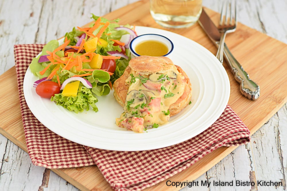 Creamed Lobster in Puffed Pastry Shell with a side green salad sits on a wooden board with a checkered napkin tucked underneath the white plate