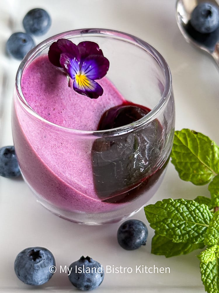 Small liqueur glass filled with panna cotta and a dollop of blueberry sauce. Garnished with a pretty purple flower and surrounded by blueberries and mint leaves