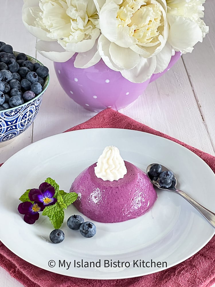 Unmolded Panna Cotta on white plate surrounded by blueberries, mint leaf, and a purple flower. White peonies in purple polka dotted vase and a blue bowl of blueberries are in background