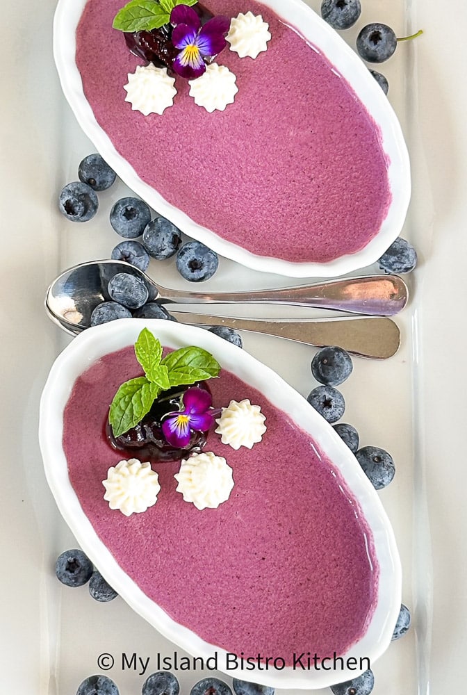 Panna Cotta garnished with tiny dollops of whipped cream, a purple flower, and mint leaves in shallow white dishes.