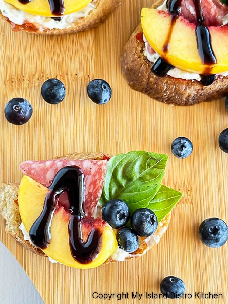 Tiny crostini topped with peach wedge, blueberries, and a cold cut. Garnished with basil leaves and balsamic glaze