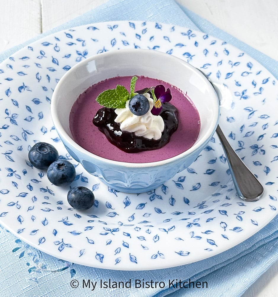 Panna Cotta topped with blueberry sauce and whipped cream in serving dish which sits on a pretty blue and white plate