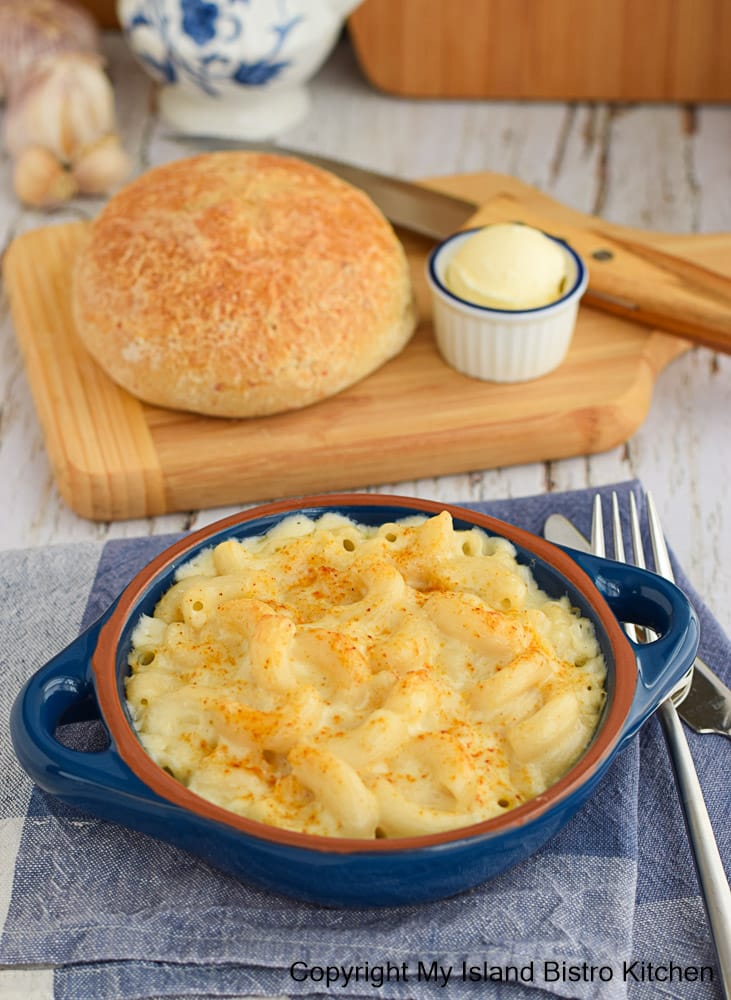 An individual serving of Mac 'n Cheese sits on blue napkin with bread and butter on a wooden board in background