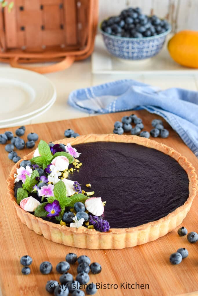 A fruit curd tart sits atop a wooden board surrounded by blueberries