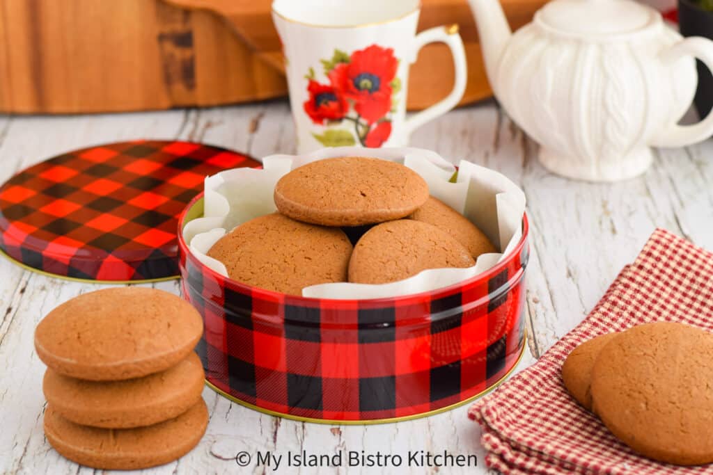 A red and black plaid tin is filled with homemade cookies and sits on counter beside a stack of the cookies. A white teapot and red and white mug appears in the background.