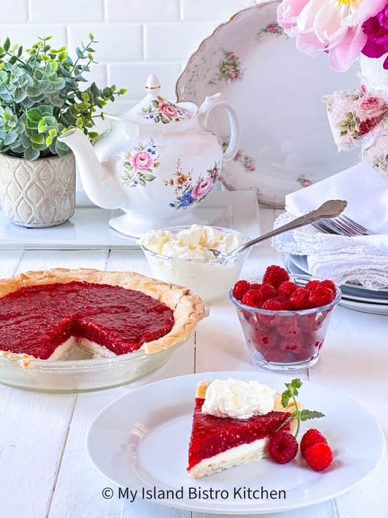 A slice of Raspberry Pie sits on a white plate with bowls of fresh raspberries and whipped cream in the background alongside the pie in the pie plate. A pretty floral teapot and plates with napkins and forks on top sit in the background.
