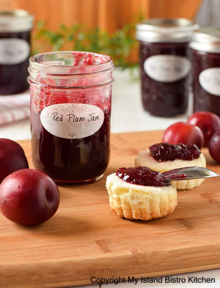 A jar of plum jam sits on a wooden board with biscuits topped with the dark red jam