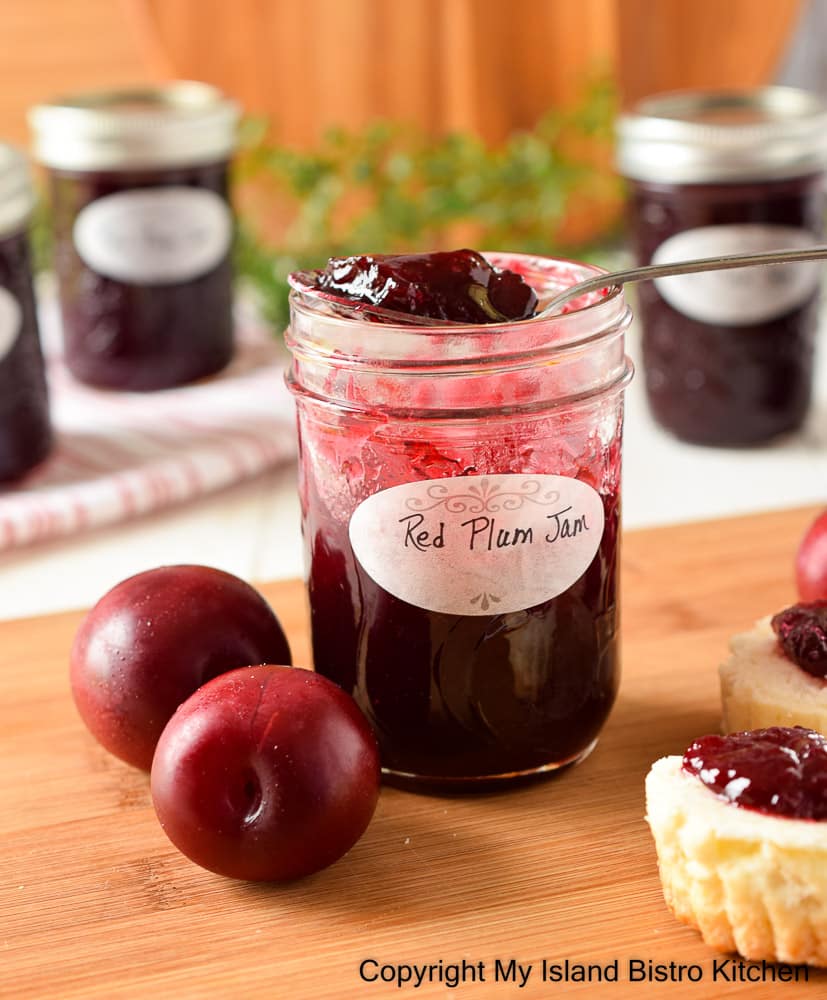 A spoonful of jam sits atop a jar of plum jam with Vampire Red Plums on one side of the jar and biscuits topped with the jam on the opposite side. Jars of jam are in the background.